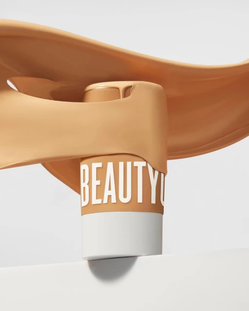Tan foundation bottle with the word 'BEAUTY' around it, displayed with pouring liquid on white, for beauty and fashion promotion.