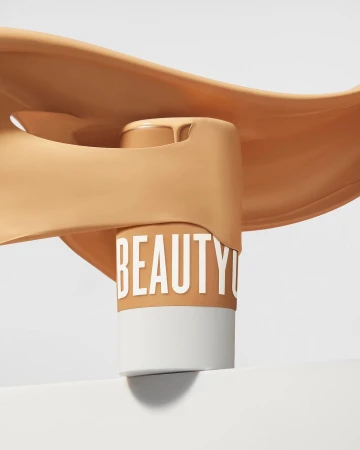 Tan foundation bottle with the word 'BEAUTY' around it, displayed with pouring liquid on white, for beauty and fashion promotion.
