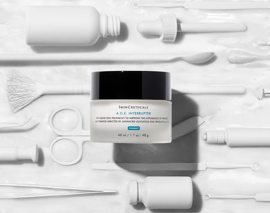 Skinceuticals A.G.E. Interrupter anti-aging cream jar is displayed on a white surface surrounded by white cosmetic tools.