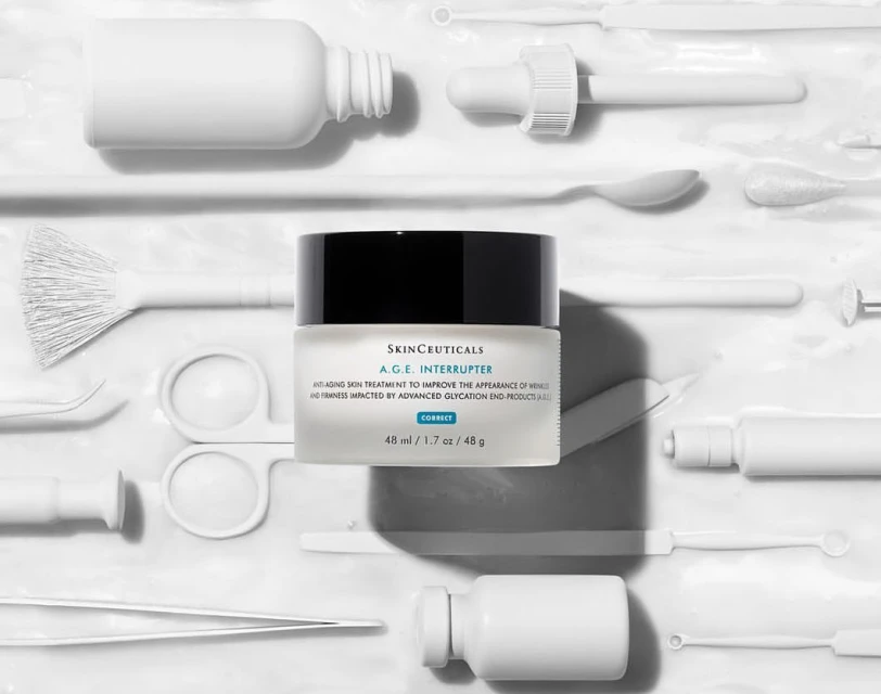 Skinceuticals A.G.E. Interrupter anti-aging cream jar is displayed on a white surface surrounded by white cosmetic tools.