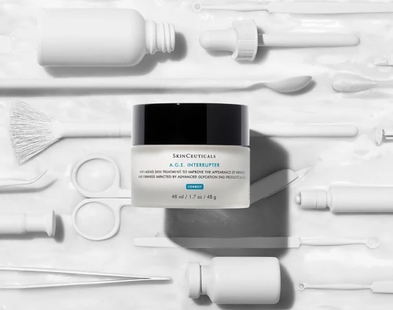 Skinceuticals A.G.E. Interrupter anti-aging cream jar is displayed on a white surface surrounded by white cosmetic tools.
