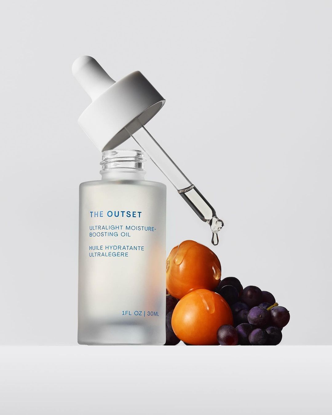 Cosmetic oil in a frosted glass bottle with dropper, featuring grapes and golden berries on a white background for beauty photography.