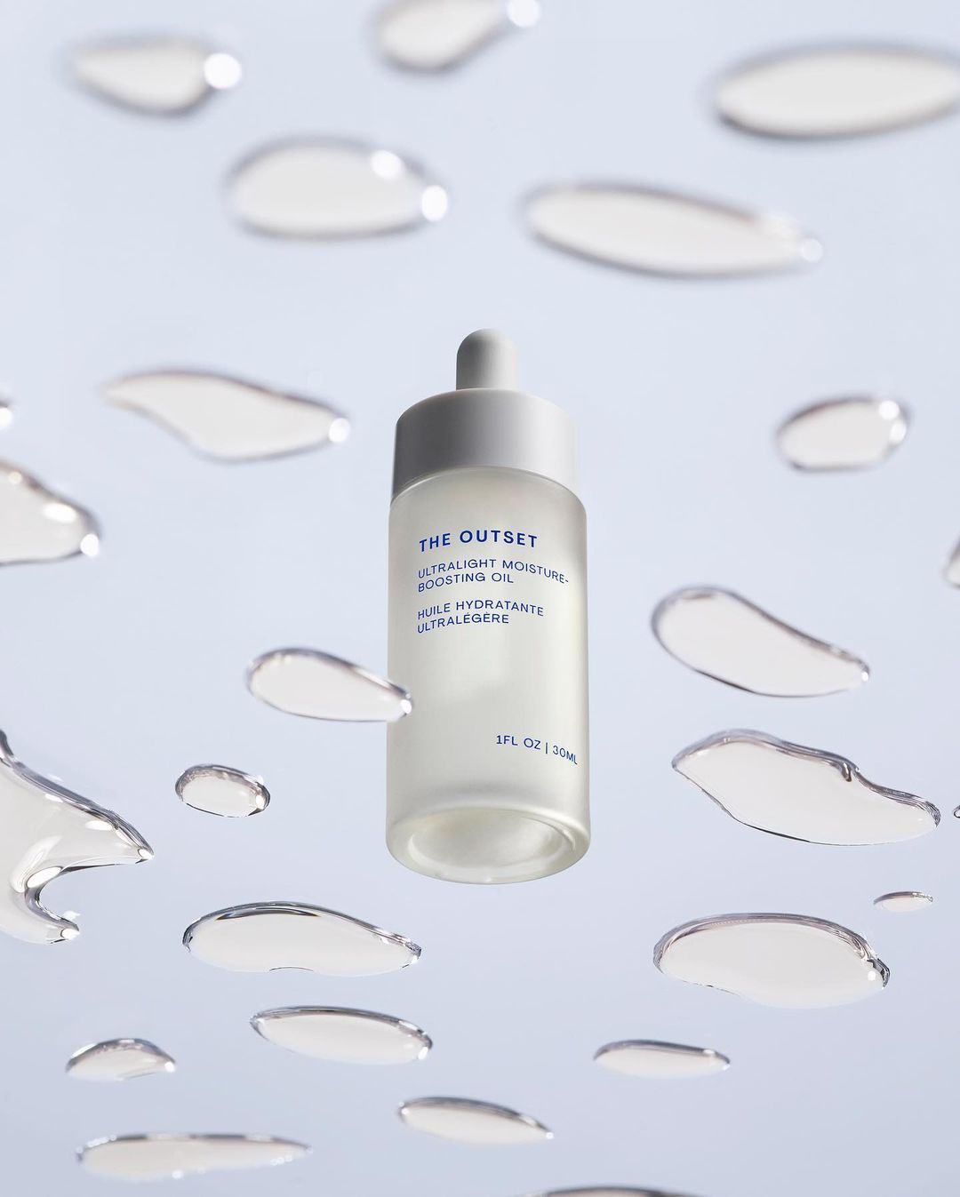 Cosmetic oil bottle with dropper, featuring The Outset brand, surrounded by water droplets on a light blue background, product shot.