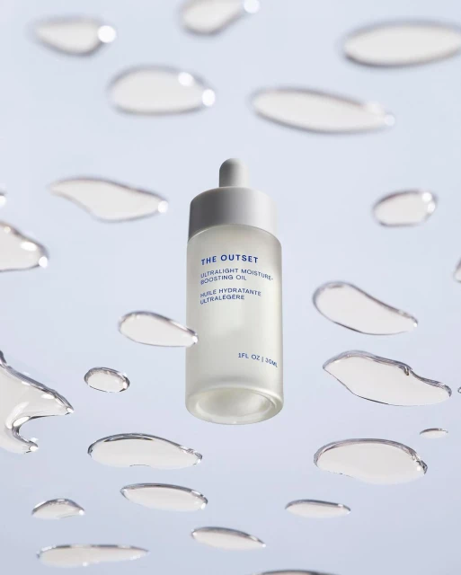 Cosmetic oil bottle with dropper, featuring The Outset brand, surrounded by water droplets on a light blue background, product shot.