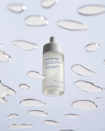 Cosmetic oil bottle with dropper, featuring The Outset brand, surrounded by water droplets on a light blue background, product shot.