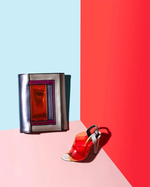 Fashion still life of a transparent heeled sandal and a color block handbag on a pink, blue, and red background in a studio.