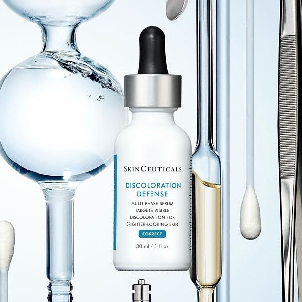 SkinCeuticals Discoloration Defense serum bottle sits in foreground, skincare product shot with lab glassware and accessories, white background.