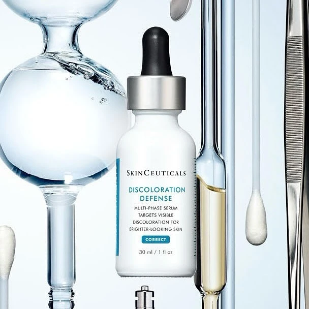 SkinCeuticals Discoloration Defense serum bottle sits in foreground, skincare product shot with lab glassware and accessories, white background.