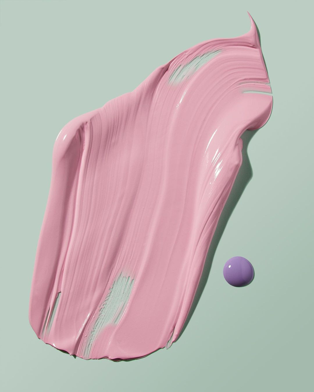 Close-up of a swipe of pink paint or cosmetic cream with a small purple sphere, against a light green background, studio lighting.