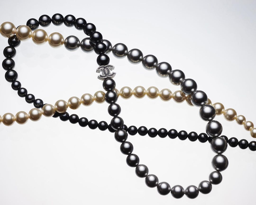 Elegant still life of Chanel pearl necklaces in black, grey, and cream shades arranged artistically on a reflective white surface.
