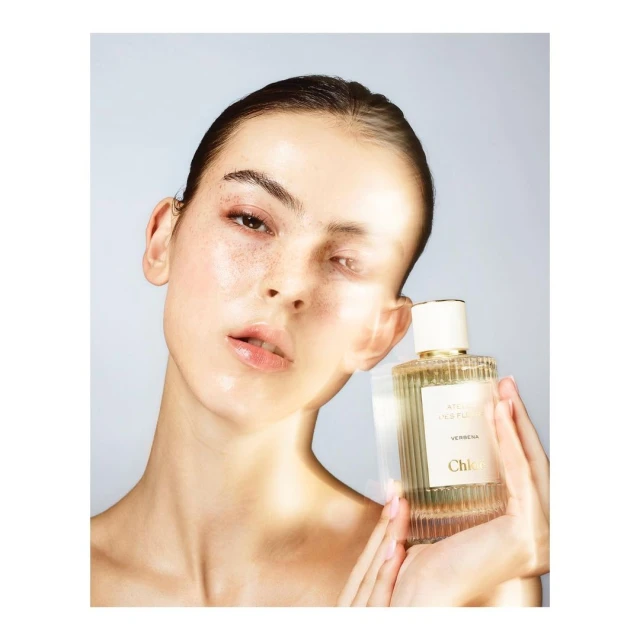 Close-up of a young adult woman holding a Chloe perfume bottle against a light blue background in a beauty and fashion shot.