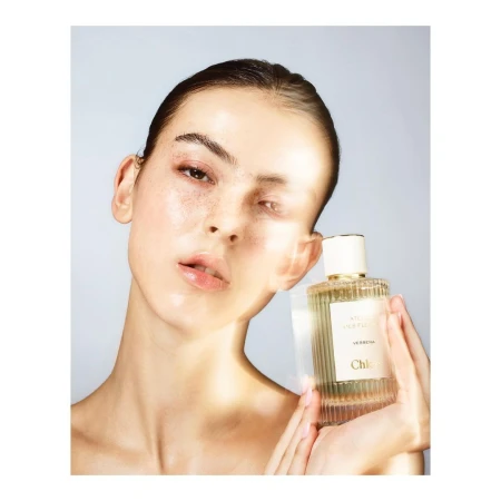 Close-up of a young adult woman holding a Chloe perfume bottle against a light blue background in a beauty and fashion shot.