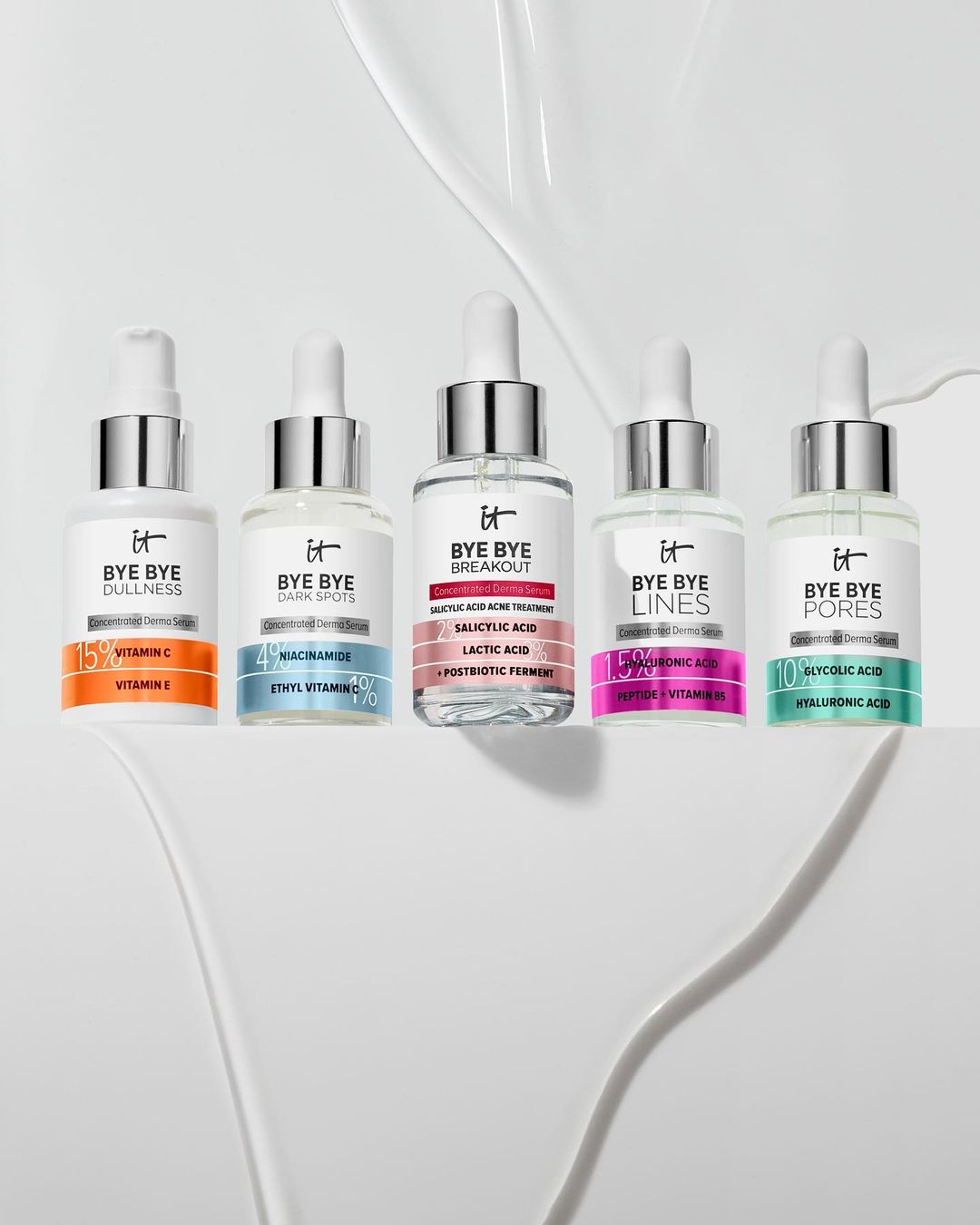 Five bottles of IT Cosmetics Bye Bye serums sit on a white surface, each targeting different skin concerns, like dullness, dark spots, and pores.