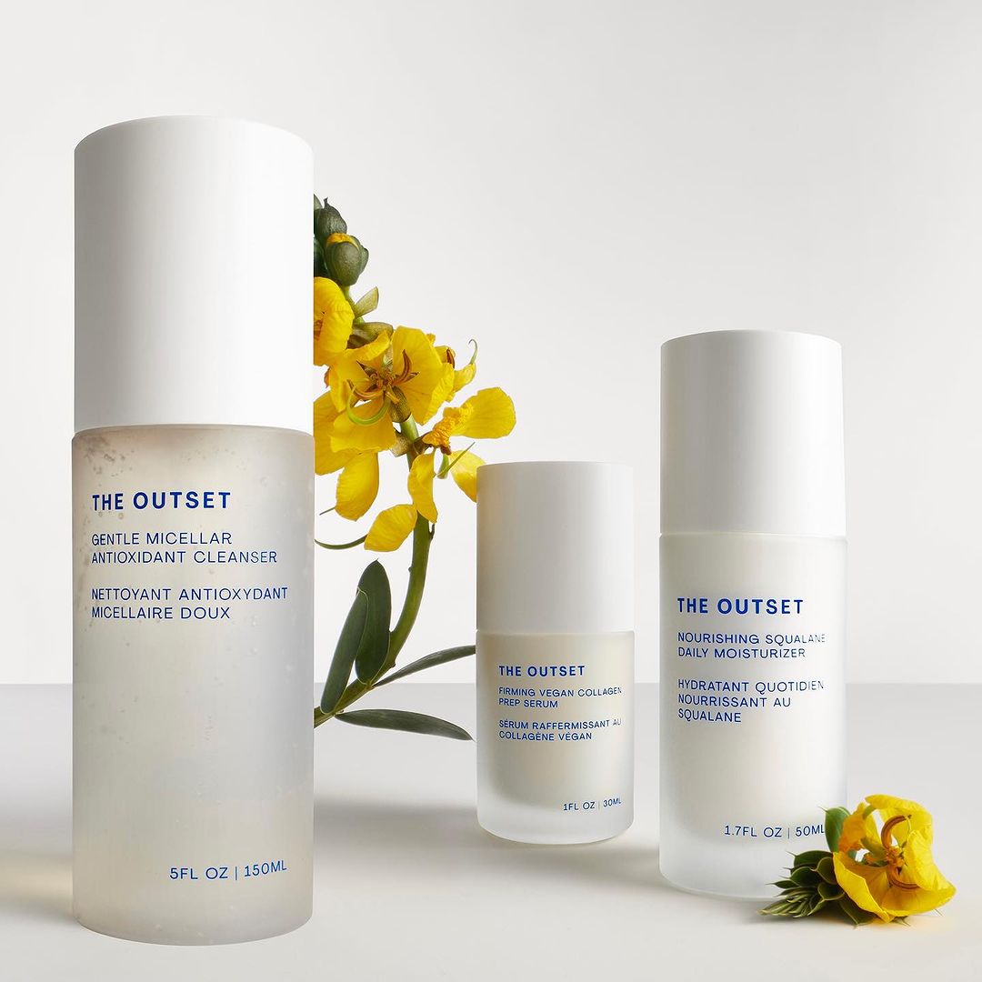 The Outset skincare product line featuring cleanser, serum, and moisturizer in frosted bottles, displayed with yellow flowers on a white surface.