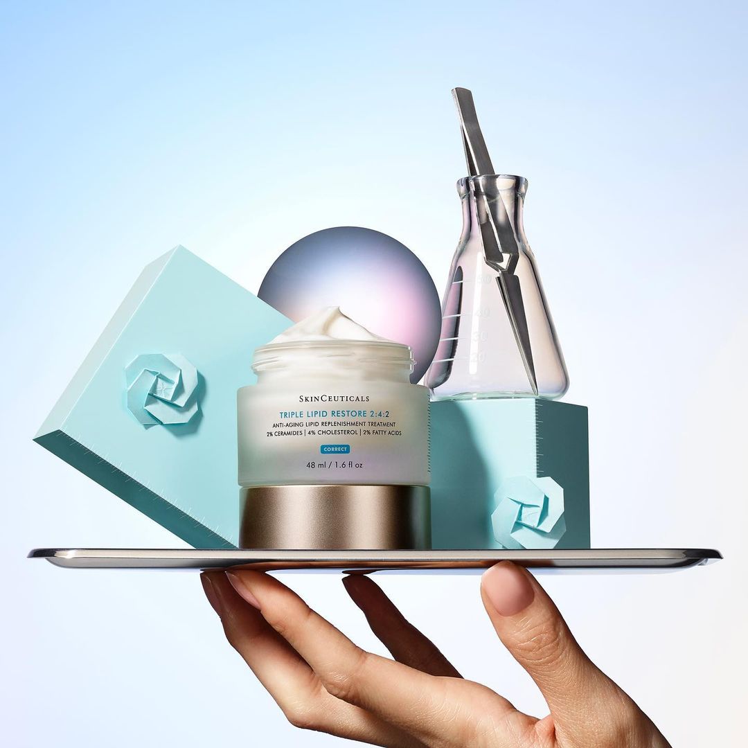 Cosmetic product presentation: skincare cream jar on a silver tray held by a hand, laboratory glassware, soft studio lighting, pastel background.
