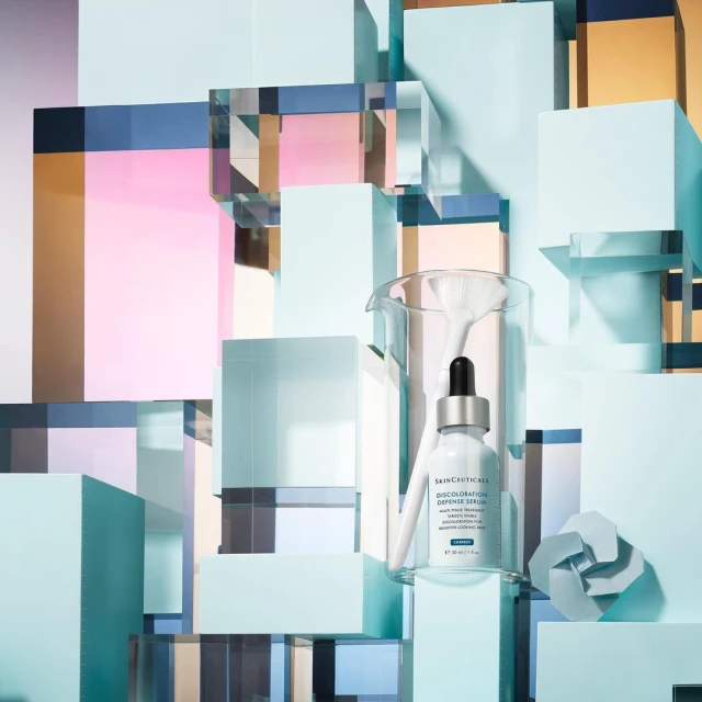 SkinCeuticals discoloration defense serum bottle with dropper presented in a beaker against a geometric background of pastel cubes.