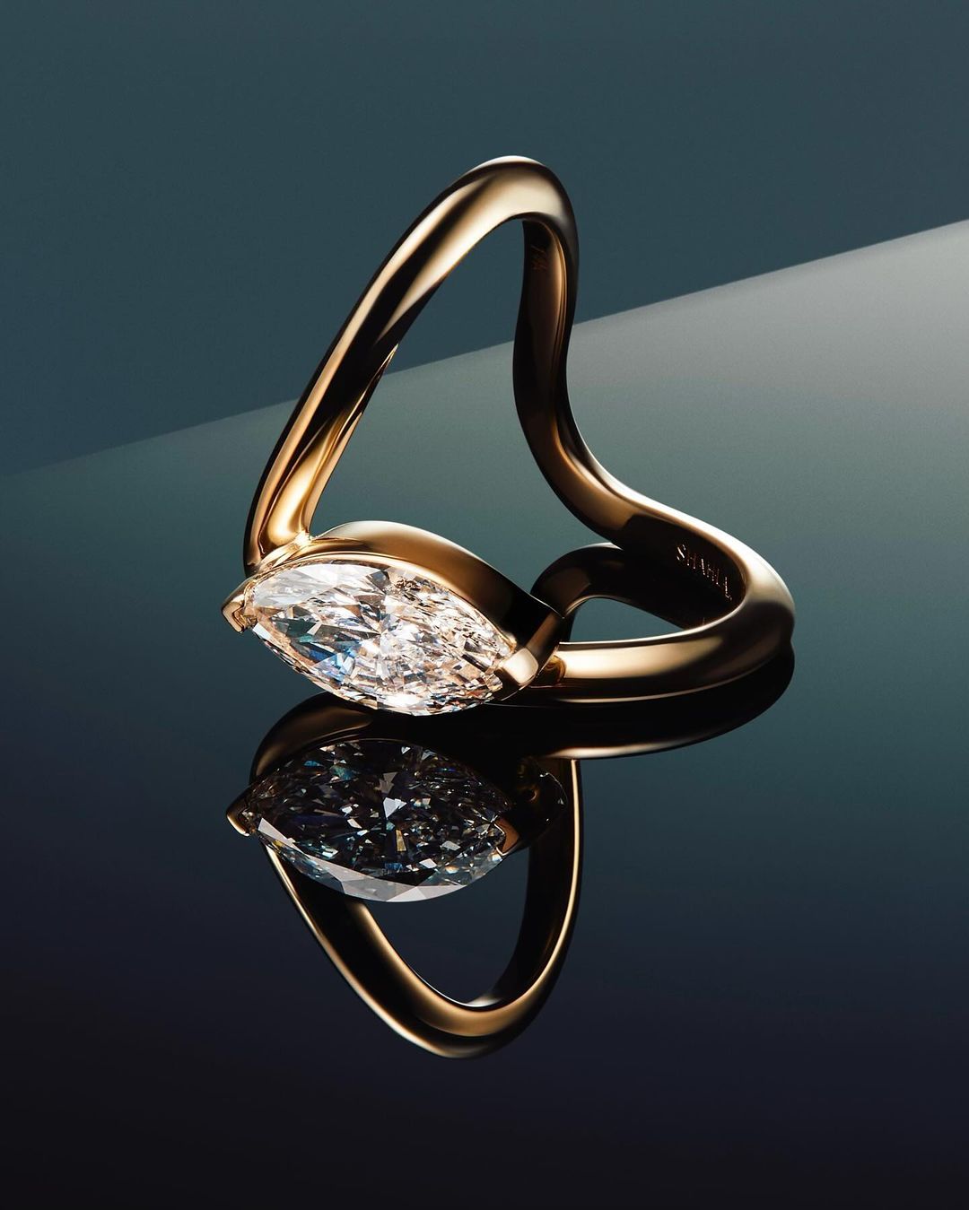 Close-up studio shot of a gold ring featuring a large, clear marquise-cut diamond, reflected on a glossy dark surface.