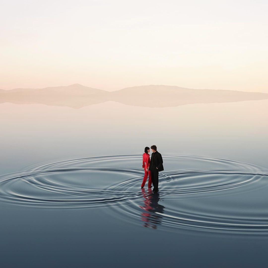 A couple is wading through a shallow body of water, creating ripples on the surface, with distant mountains under a pastel sky in the background.