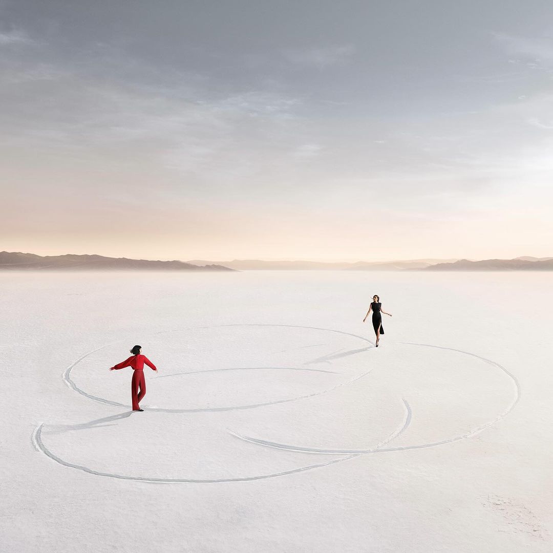 A woman in a black dress walks across a white expanse toward the camera; another woman in a red jumpsuit stands in the distance.