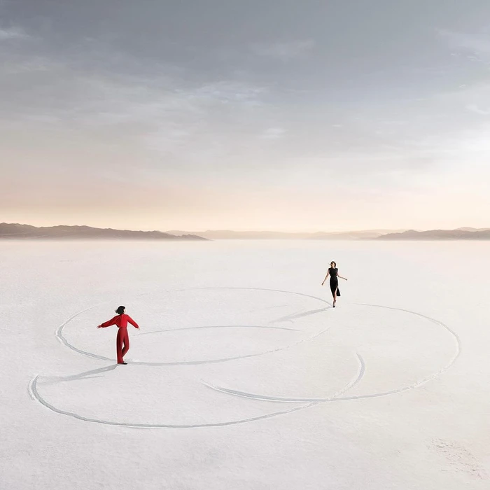 Two figures standing on a vast white salt flat beneath a pale hazy sky, with large circular track lines inscribed in the ground.