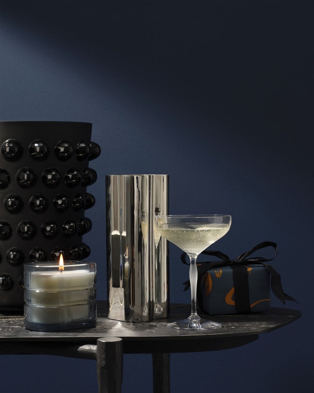 A lit candle, cocktail glass with bubbly drink, wrapped gift, and a vase on a dark metal table, against a dark blue background.