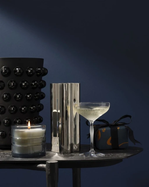 A lit candle, cocktail glass with bubbly drink, wrapped gift, and a vase on a dark metal table, against a dark blue background.