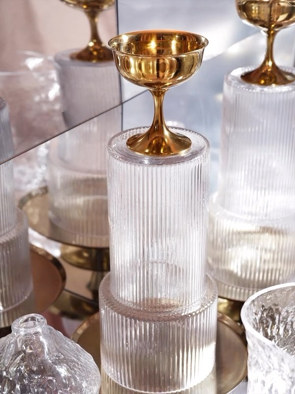 Close-up of a gold coupe glass sitting on top of textured clear glass pedestal with mirror background, studio lighting, and neutral color palette.