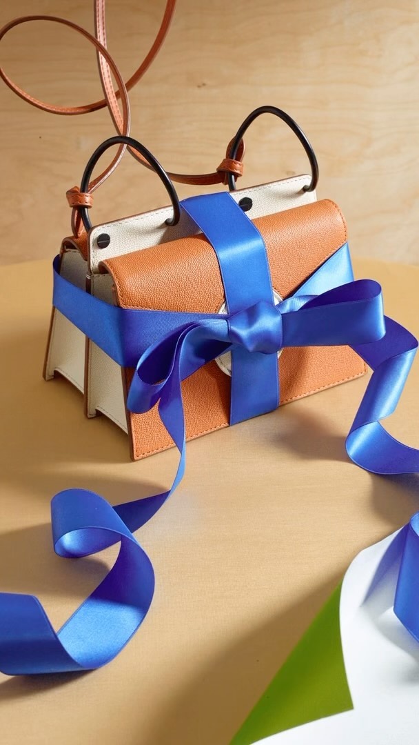 Tan leather handbag wrapped in a blue ribbon on a beige surface, with a light brown wooden background creating a minimalist fashion image.