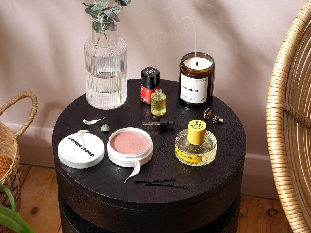 Cosmetic and self-care products are displayed on a black side table next to a wicker chair, with a light pink wall in the background.