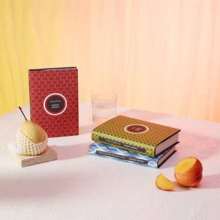 Still life shot of three George Orwell books, a pear in protective wrap, a drinking glass, and a halved peach on a white surface.