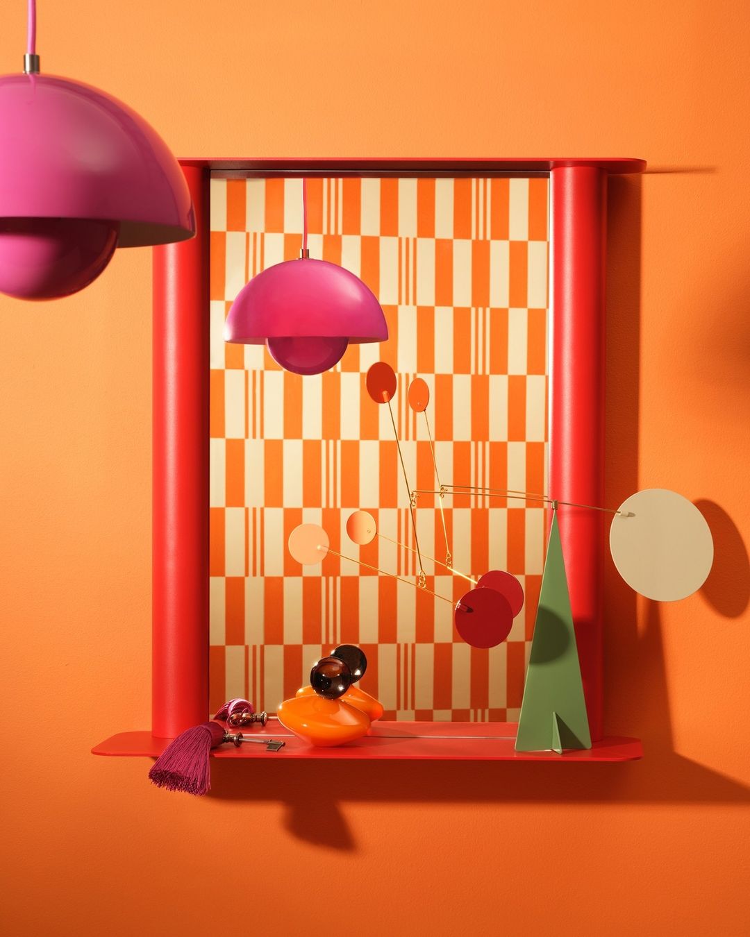 Still life composition features pink pendant lamps, red window frame, geometric wallpaper, decorative objects, and orange wall.
