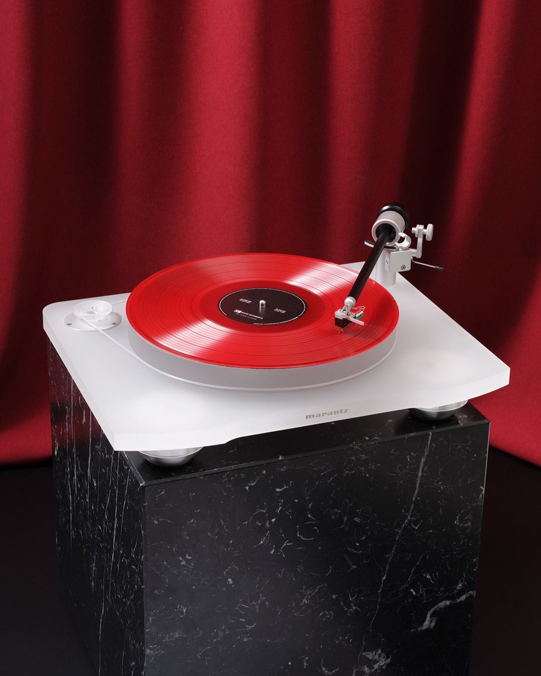 A white Marantz turntable with a red vinyl record sits atop a black marble stand, set against a blurred red curtain backdrop.