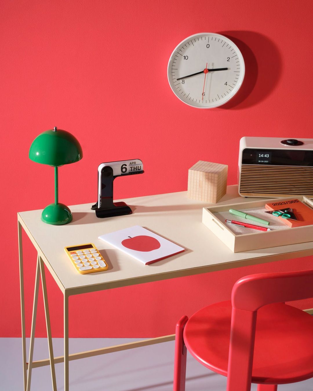 Still life of a desk with a yellow calculator, calendar, lamp, clock, and other objects against a red wall and gray floor.