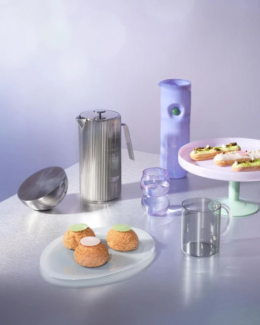 Still life featuring choux pastries, clear glass mugs and cake stand against soft blue background with a reflective surface. Food photography.