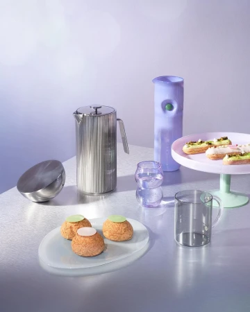 Still life featuring choux pastries, clear glass mugs and cake stand against soft blue background with a reflective surface. Food photography.