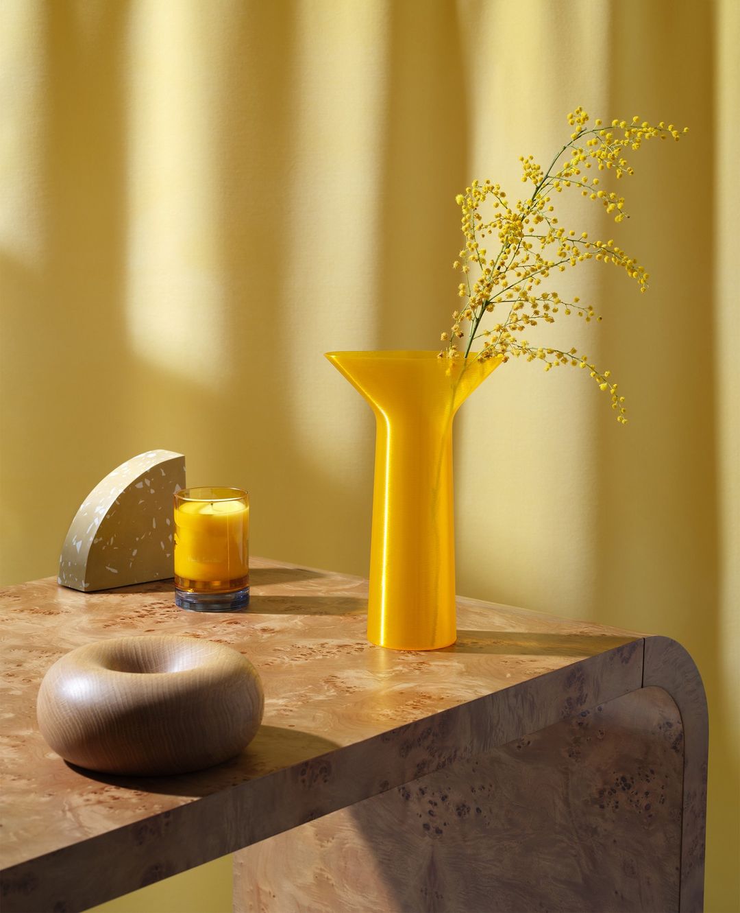 Yellow-toned still life featuring a vase with mimosa, a candle, and geometric objects on a marble table against a draped backdrop.
