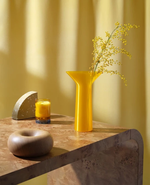Yellow-toned still life featuring a vase with mimosa, a candle, and geometric objects on a marble table against a draped backdrop.