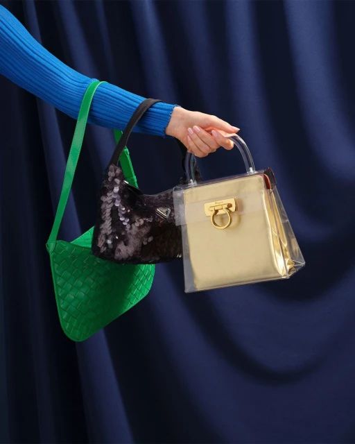 Close-up shot of three handbags with different textures and materials, held by a woman's hand against a navy blue draped background.