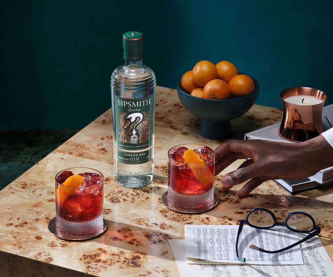 Two cocktails with a bottle of gin, oranges, sheet music, and eyeglasses on a light wood table against a dark teal background.