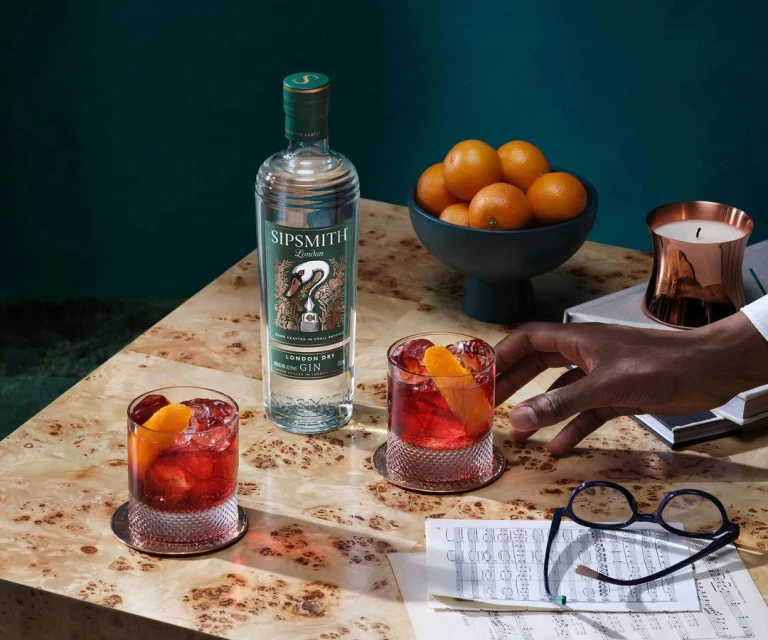 Two cocktails with a bottle of gin, oranges, sheet music, and eyeglasses on a light wood table against a dark teal background.