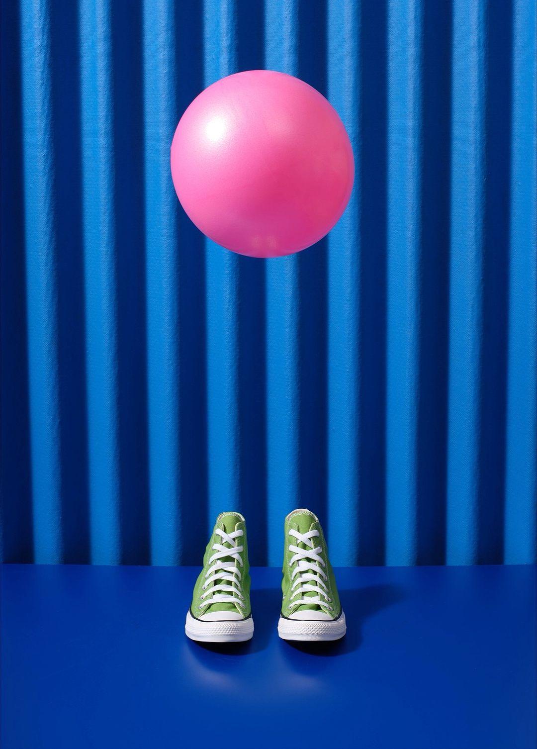 Two green high-top sneakers sit on a blue surface beneath a pink ball against a striped blue background, creating a vibrant and playful studio shot.