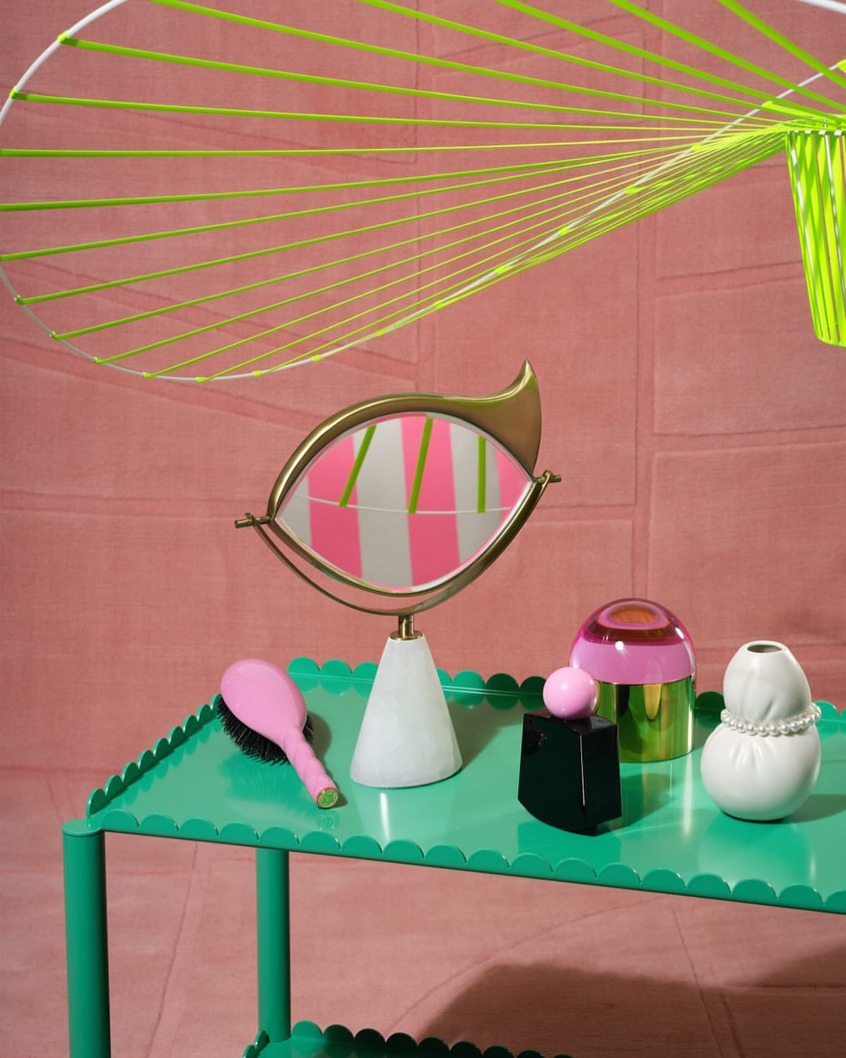 Still life composition featuring a mirror, cosmetics, a brush, and decorative elements on a green table against a pink backdrop.
