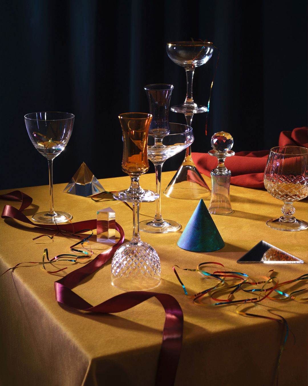 Still life photo of multiple stemmed glasses arranged on a table with colorful geometric shapes, ribbons and a dark curtain background.