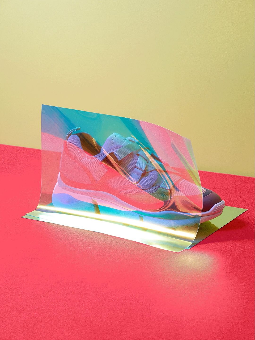 Still life of a pink and blue sneaker partially obscured by a sheet of iridescent cellophane on a red and yellow surface.