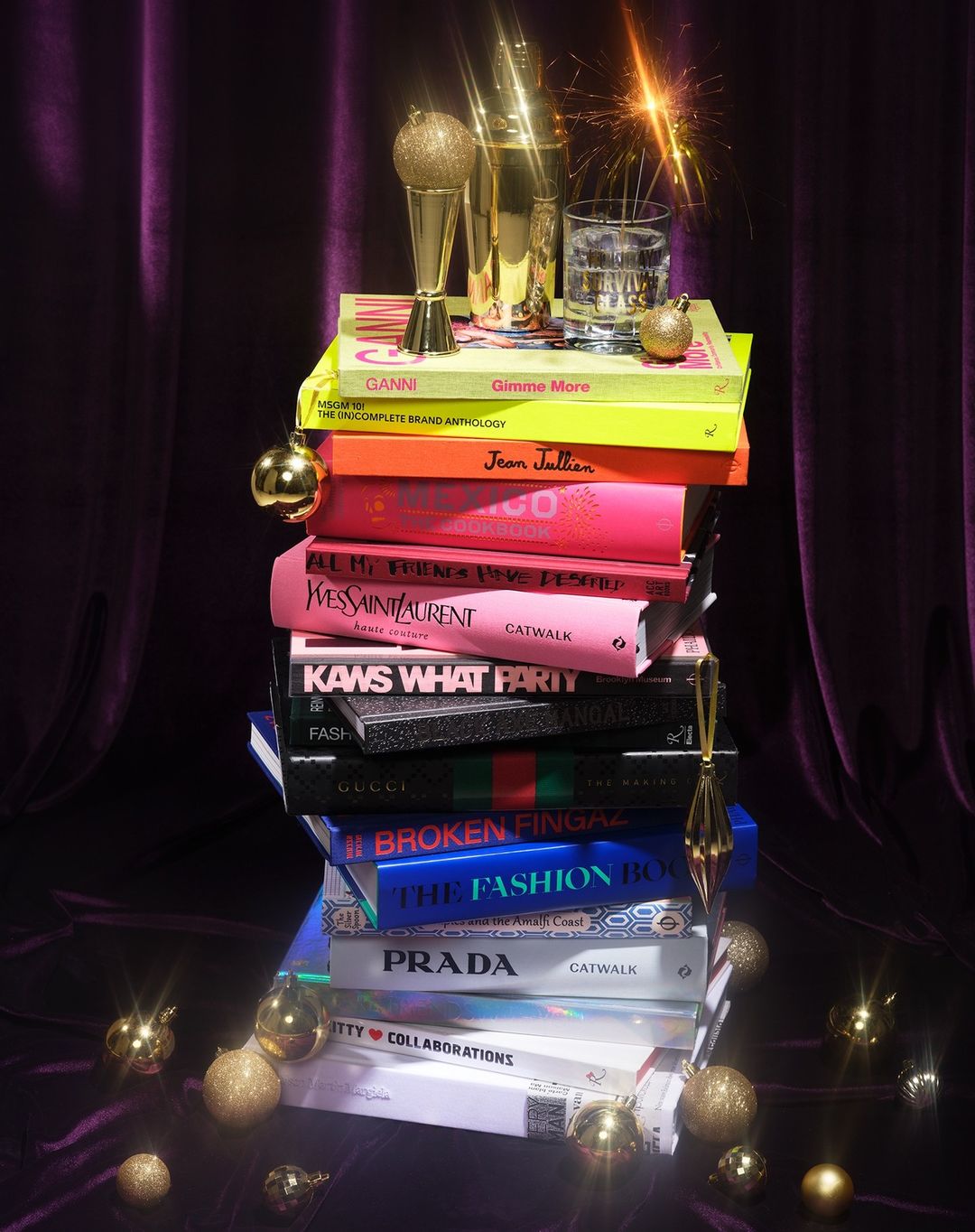 Still life of a colorful stack of fashion and art books topped with holiday ornaments, glassware, and golden cocktail shaker with sparklers.