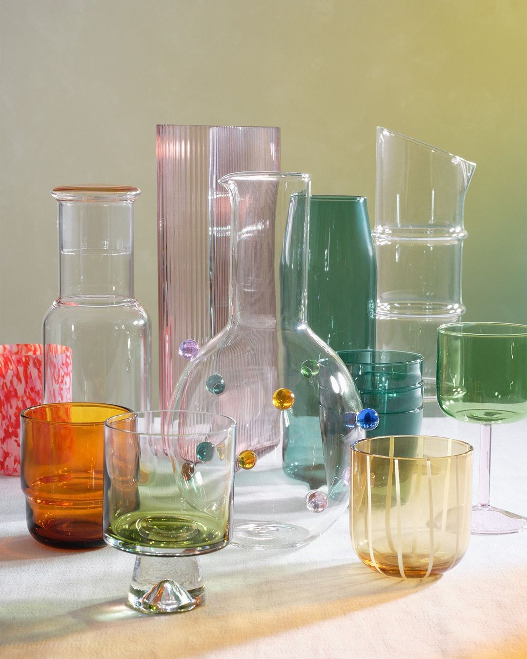 Still life of colorful glassware including carafes, glasses, and pitchers in amber, green, pink, and clear glass against a gradient background.