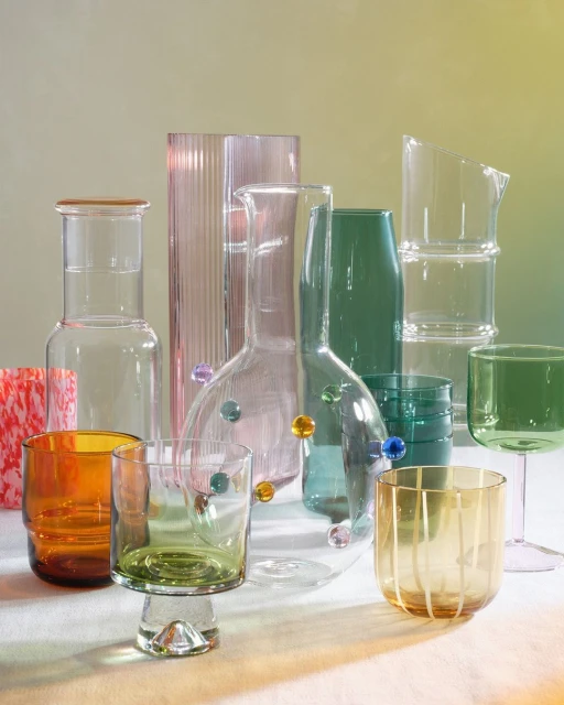 Still life of colorful glassware including carafes, glasses, and pitchers in amber, green, pink, and clear glass against a gradient background.