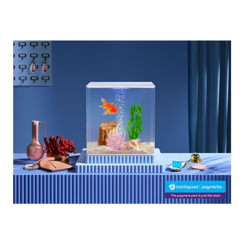 Orange goldfish swimming in a square aquarium with decorative plants, set atop a blue corrugated pedestal next to vintage props.