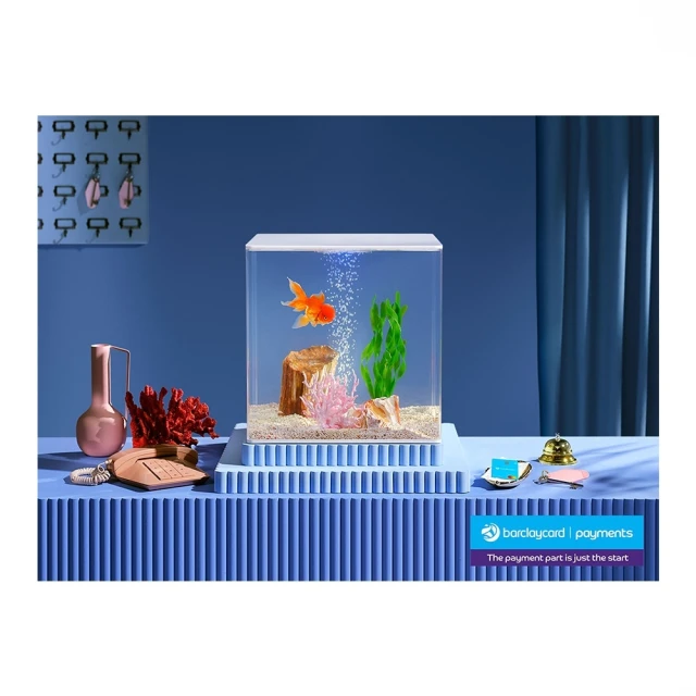 Orange goldfish swimming in a square aquarium with decorative plants, set atop a blue corrugated pedestal next to vintage props.