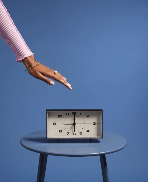 Close-up of a hand in a pink sweater over a vintage black-and-white clock sitting on a blue table against a solid blue backdrop.